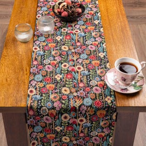 May include: A black table runner featuring a dense, colourful woven pattern of various stylised flowers in shades of red, pink, blue, yellow, and green. The runner is displayed on a wooden table with a clear glass candle holder and a floral teacup.