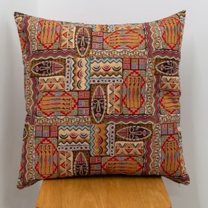 May include: Square cushion with a vibrant tribal design. The pattern includes masks and geometric shapes in brown, red, orange, and blue. The cushion is placed on a wooden stool, showcasing its intricate details and colours.