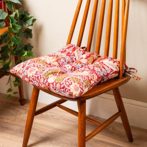 Puede incluir: Un cojín de silla con estampado floral rojo y verde con cierre de corbata. El cojín está colocado en una silla de madera.