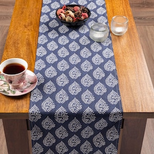 May include: A navy blue table runner featuring a repeating white leaf pattern, displayed on a wooden table with a floral teacup and saucer, a black bowl filled with decorative pinecones and berries, and two clear glass candle holders.