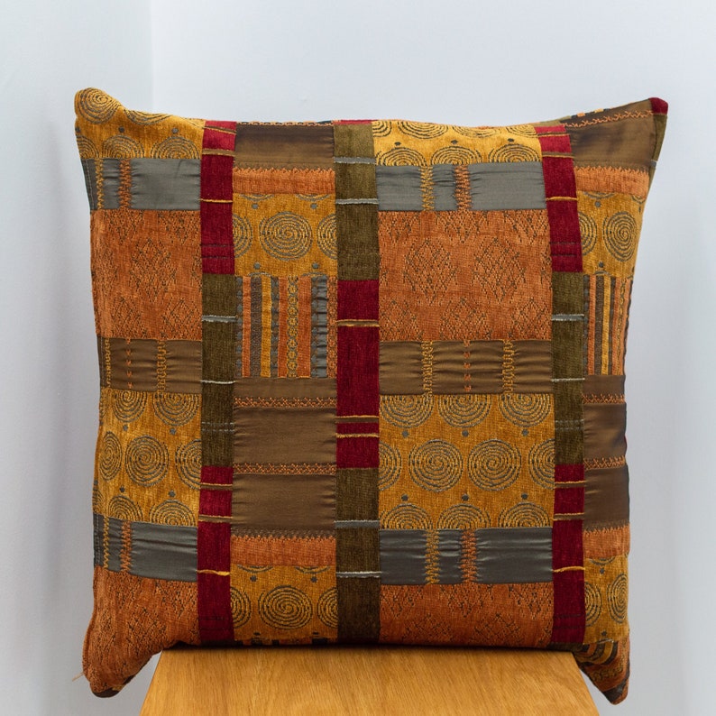 May include: A decorative throw pillow with a patchwork design featuring various shades of brown, gold, and red. The pillow has a geometric pattern with a subtle swirl design.