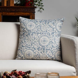 Peut inclure: Un coussin carré décoratif avec un motif floral bleu et blanc. Le coussin est posé sur un canapé de couleur claire. Le motif comprend des cercles et des motifs de feuilles, créant une esthétique vintage. Le coussin ajoute une touche d'élégance.