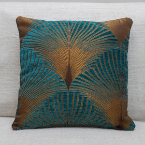 Pode incluir: Travesseiro decorativo azul-esverdeado e dourado com um padrão geométrico de penas de pavão estilizadas.