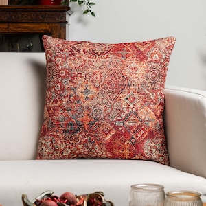 May include: A square cushion featuring a detailed, ornate pattern in shades of red, orange, and blue. The cushion is placed on a white sofa, highlighting its intricate design and texture. The cushion's design has a vintage, rug-like appearance.