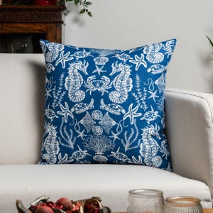 Puede incluir: Una almohada azul con un patrón de vida marina blanco. El patrón incluye caballitos de mar, estrellas de mar, conchas marinas y coral.