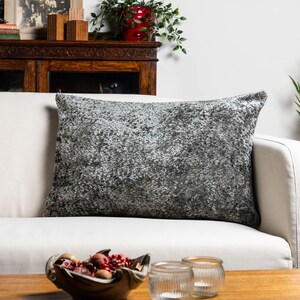 Puede incluir: Un cojín de sofá rectangular gris y blanco texturizado, colocado en un sofá blanco. El cojín es suave y esponjoso.