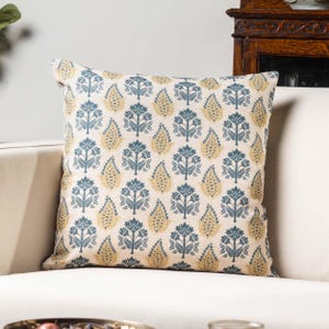 Op de afbeelding: Een decoratief kussen met een witte achtergrond en een herhalend patroon van blauwe en gele bloemenmotieven.