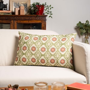 May include: A rectangular cushion with a floral design. The cushion has a light green backdrop with a repeating pattern of white and coral-coloured flowers. The cushion is placed on a white sofa.