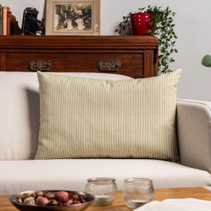 Op de afbeelding: Een rechthoekig kussen met een crème- en olijfgroen streepjespatroon. Het kussen ligt op een witte bank. De achtergrond omvat een houten dressoir, een ingelijste foto en decoratieve items.