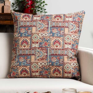 May include: Square cushion with a patchwork design. The cushion features a pattern of squares in shades of blue, red, and beige. The design includes floral and paisley motifs, creating a vintage or bohemian aesthetic.