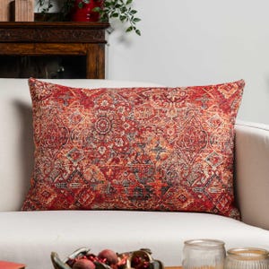 Può includere: Cuscino decorativo rettangolare con un motivo dettagliato e ornato nei toni del rosso, arancione e blu. Il cuscino è posto su un divano bianco, mettendo in risalto il suo design intricato e la sua tavolozza di colori. Il design del cuscino ricorda un tappeto tradizionale.