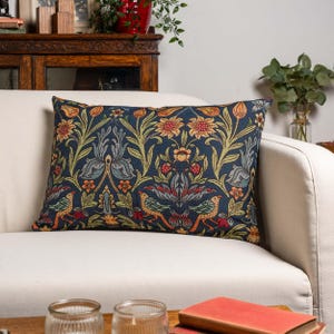 Può includere: Un cuscino decorativo rettangolare con sfondo blu navy e motivo floreale e di uccelli. Il design presenta fiori nei toni dell'arancione, giallo e rosso, con foglie verdi e uccelli stilizzati. Il cuscino è posto su un divano bianco.