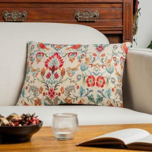 Può includere: Un cuscino rettangolare con una base in tessuto color crema e un motivo a arazzo intrecciato. Il design presenta motivi floreali simmetrici, viticci e piccoli uccelli nelle tonalità del rosso, blu, arancione e verde tenue.