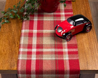 May include: A red and black toy car sits on a red, beige, and white plaid table runner. A red pot with a green plant is in the background. The table is made of wood.