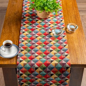 Puede incluir: Un camino de mesa con estampado geométrico colorido con una variedad de colores, incluyendo rojo, azul, verde y amarillo. El camino de mesa está colocado sobre una mesa de madera, con una pequeña planta en una cesta tejida, dos candelabros de vidrio y una taza de café.