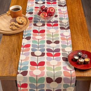 Op de afbeelding: Een kleurrijke tafelloper met bloemenpatroon, met fruit en koekjes op een houten tafel. Het patroon toont gestileerde bloemen in rood, roze, blauw en paars. Een kopje koffie staat op een houten dienblad.