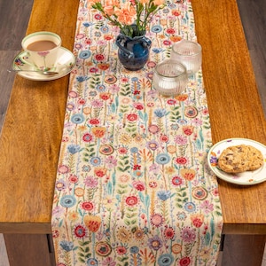 Op de afbeelding: Een bloemen tafelloper met een crèmekleurige achtergrond en een kleurrijk bloemenpatroon. De tafel is gedekt met een kop en schotel, een vaas met bloemen, twee glazen kandelaars en een bord met een koekje.