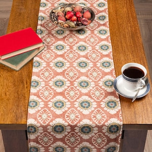 Peut inclure: Chemin de table à motifs floraux dans les tons corail, blanc et bleu, avec un bol de pot-pourri, une tasse de café et des livres sur une table en bois. Le motif floral est répété.