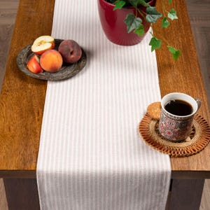 Könnte beinhalten: Ein Holztisch mit einem hellrosa und weiß gestreiften Tischläufer. Ein dekorativer Teller enthält geschnittene Äpfel und Pfirsiche. Eine Tasse Kaffee steht auf einem dekorativen Untersetzer, neben einem Keks. Ein roter Topf mit einer grünen Pflanze steht ebenfalls auf dem Tisch.