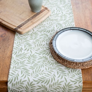 Peut inclure: Un chemin de table vert et blanc avec un motif de feuilles. Le chemin de table est sur une table en bois avec une assiette et une planche à découper en bois.