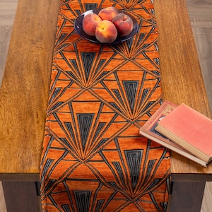 May include: An orange Art Deco table runner with a geometric fan design in black and grey. The runner is on a wooden table, with a small plate of peaches and two books.