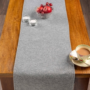 Op de afbeelding: Een grijze visgraat tafelloper ligt op een houten tafel. Een glazen kom met rode appels en druiven staat op de loper, samen met twee kleine kaarshouders en een theekopje.