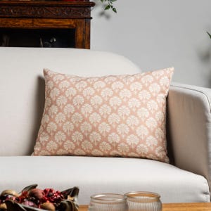 May include: A rectangular cushion featuring a repeating floral design in cream on a peach-toned fabric. The cushion is placed on a white sofa. The design adds a touch of elegance to the living room.
