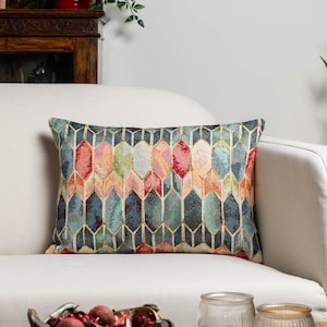 Pode incluir: Uma almofada retangular decorativa com um padrão geométrico em tons de azul, verde-azulado, rosa, laranja e verde. A almofada está sobre um sofá branco. O design apresenta um padrão repetitivo de formas alongadas e conectadas.