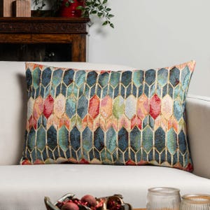 Pode incluir: Almofada decorativa retangular com um padrão geométrico em tons de azul, vermelho, laranja e verde. A almofada está colocada num sofá branco, adicionando um toque de cor à decoração.