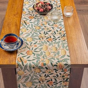 May include: A woven table runner featuring a repeating pattern of yellow lemons, orange fruits, and green leaves on a cream background. A blue and white patterned teacup on a saucer, a glass bowl of potpourri, and a clear glass candle holder sit on top.