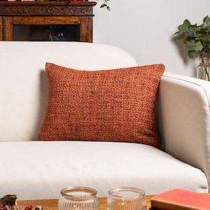 Könnte beinhalten: Ein orangefarbenes rechteckiges Kissen mit gewebter Textur liegt auf einem weißen Sofa. Die Farbe des Kissens ist ein warmer, erdiger Ton, und das Gewebe erzeugt ein subtiles Muster. Das Sofa ist Teil eines Wohnzimmers.