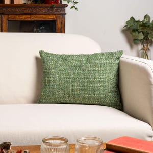Puede incluir: Un cojín rectangular verde tejido descansa sobre un sofá blanco. El cojín tiene un aspecto texturizado con diferentes tonos de verde. El sofá está en un salón, con un mueble de madera y objetos decorativos en el fondo.