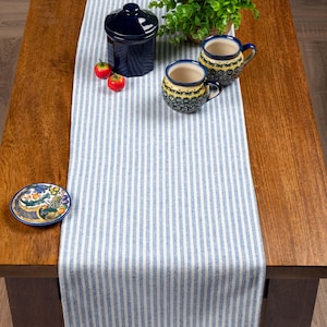 May include: A wooden table features a blue and white striped table runner. Two decorative mugs with blue handles and a small ceramic dish are on the table. A dark blue jar with a lid and two small red tomatoes are also present.