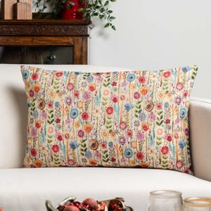 May include: Rectangular cushion with a floral design. The cushion has a cream-coloured background with a colourful array of embroidered flowers in shades of red, blue, yellow, and purple. The cushion is placed on a white sofa.