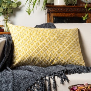 Può includere: Un cuscino giallo con un motivo geometrico bianco. Il cuscino è su un divano bianco con una coperta grigia.