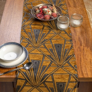 Può includere: Una tovaglia color giallo senape con un motivo geometrico a ventaglio grigio e nero. Una ciotola d'argento con oggetti decorativi, due portacandele in vetro, una ciotola, un piatto e un cucchiaio sono sul tavolo di legno.