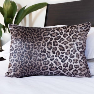 Op de afbeelding: Een rechthoekig decoratief kussen met een luipaardprint in bruin, grijs en zwart. Het kussen ligt op een wit bed, met een donkerbruin hoofdbord en een groene plant op de achtergrond.