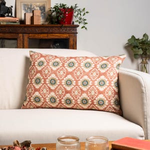 May include: A rectangular cushion with a floral pattern in shades of rust, cream, blue, and yellow. The cushion is placed on a white sofa, adding a touch of colour and pattern to the neutral backdrop. The design features a repeating floral motif.