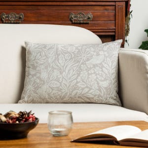 Può includere: Un cuscino rettangolare caratterizzato da un motivo botanico color beige chiaro e bianco. Il design presenta intricate illustrazioni di vari fiori, foglie e piccoli uccelli intrecciati su tutta la superficie del rivestimento in tessuto.