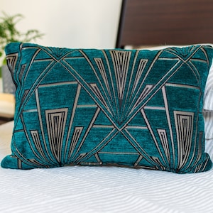 May include: Teal rectangular decorative pillow with a geometric art deco design in silver and black. The pillow is made of a textured fabric and rests on a white and gray striped bedspread. The pillow is approximately 12 inches by 20 inches.