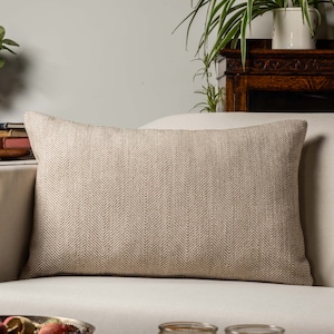 Op de afbeelding: Een beige kussen met een herringbone patroon op een witte bank.