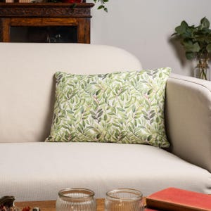 Puede incluir: Un cojín decorativo rectangular con un estampado botánico de hojas en tonos verdes, grises y rosas. El cojín está colocado sobre un sofá color crema. El diseño del cojín presenta un patrón repetido de hojas y tallos.