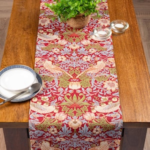 Puede incluir: Un camino de mesa rojo con un estampado floral y de pájaros. El camino de mesa presenta un diseño repetido de pájaros, flores y hojas en tonos crema, verde y azul. Una pequeña planta en maceta, un cuenco y velas están sobre la mesa de madera.