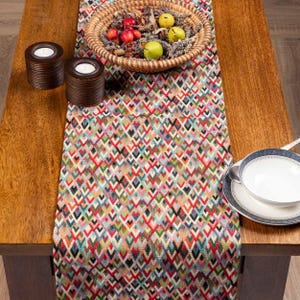 Könnte beinhalten: Ein Holztisch mit einem gemusterten Tischläufer. Der Läufer hat ein geometrisches Design in Rot, Grün, Blau und Gelb. Ein Weidenkorb mit Obst und Kerzen steht auf dem Tisch.