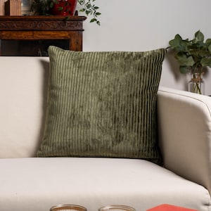May include: An olive green corduroy cushion with a vertical ribbed design sits on a cream-coloured sofa. The sofa has a curved arm. The background features a wooden cabinet and some greenery.