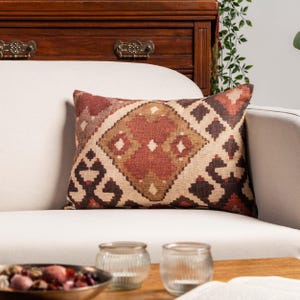 May include: A rectangular cushion with a geometric pattern in red, brown, and beige tones. The cushion is placed on a white sofa, with a wooden cabinet in the background. The design features diamond and square shapes. The cushion is likely made of fabric.