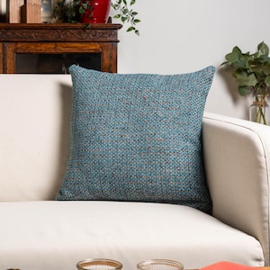 Op de afbeelding: Een vierkant sierkussen in tinten van blauwgroen, blauw en bruin. Het kussen ligt op een witte bank. De geweven stof heeft een subtiele, gevlekte uitstraling, wat zorgt voor visuele interesse. Een decoratief element voor in huis.