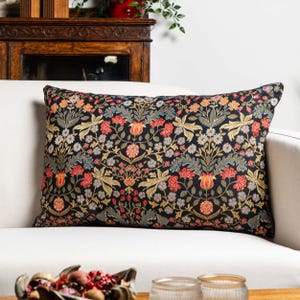 Può includere: Un cuscino decorativo rettangolare con un motivo floreale. Il cuscino presenta uno sfondo nero con un motivo ripetuto di fiori e foglie rosse, arancioni, blu e dorate. Il cuscino è appoggiato su un divano bianco.