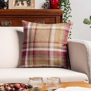 Può includere: Cuscino decorativo quadrato con motivo a quadri nei toni del beige, bordeaux e bianco. Il cuscino è su un divano bianco, con un tavolo in legno e oggetti decorativi in primo piano.