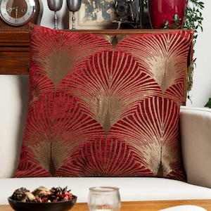 Op de afbeelding: Een vierkant sierkussen van dieprode, fluweelachtige stof met een herhalend goudkleurig, metallic waaierpatroon. Het ontwerp bestaat uit stralende lijnen die symmetrische, bladachtige motieven vormen over het gehele oppervlak van het kussen.